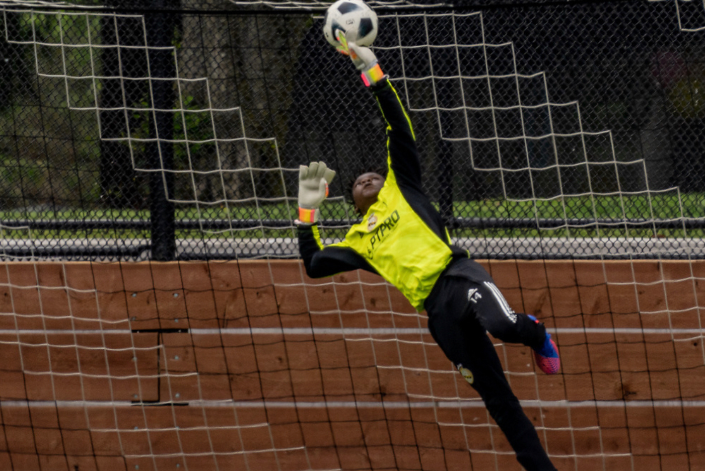 Male soccer goalkeeper in Black and yellow uniform diving to save a soccer ball from under the crossbar of the goal. 